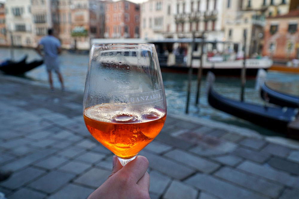 A Glass of Aperol Spritz with a blurry view of the canal in the background in Venice, Italy. 