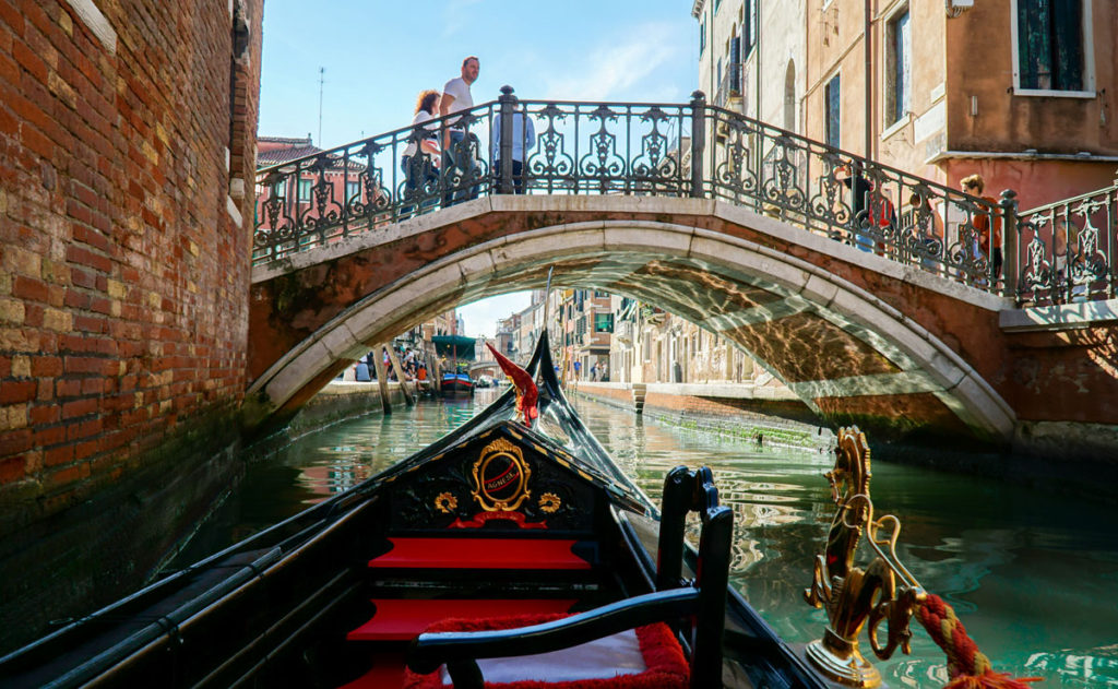 Riding the gondola through a canal under the stone bridges of Venice, Italy - Kraska Fox